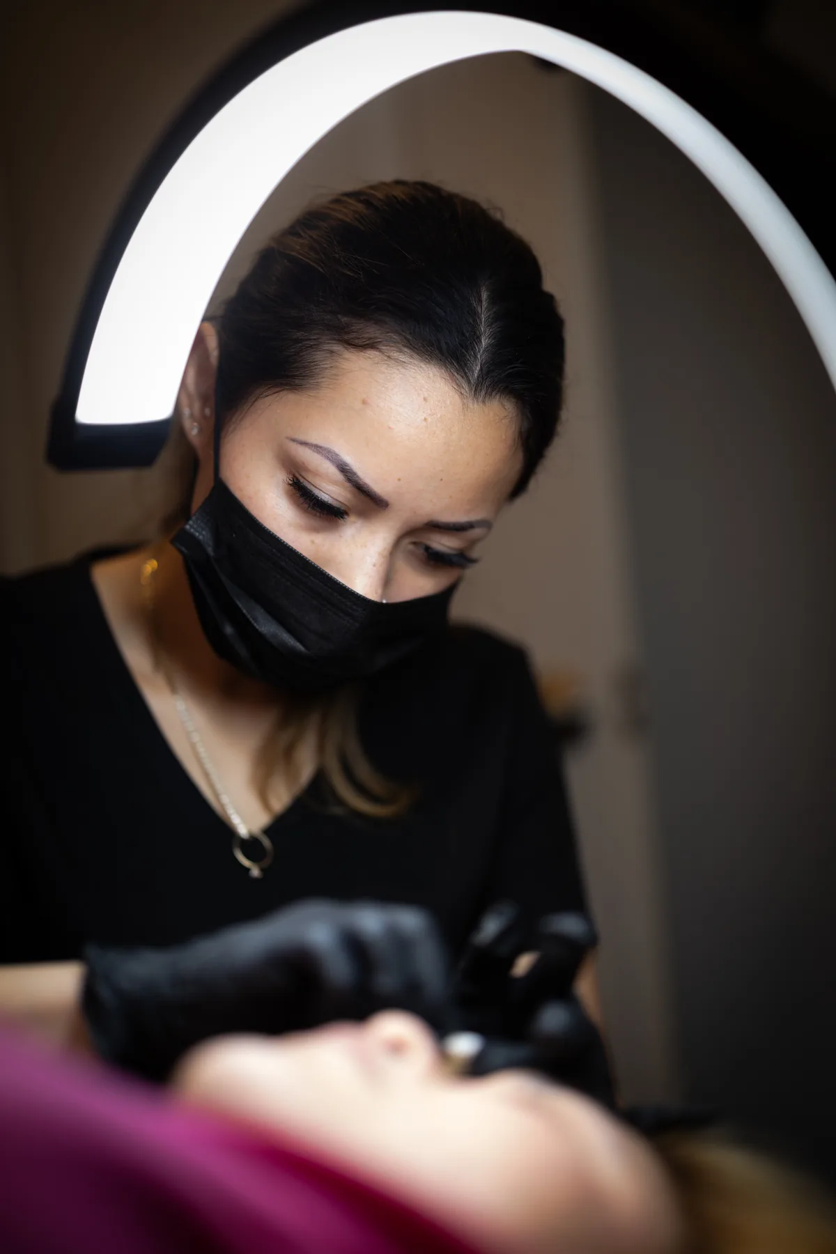 Sheyla working under a ring light, masked, focused on a client's brow